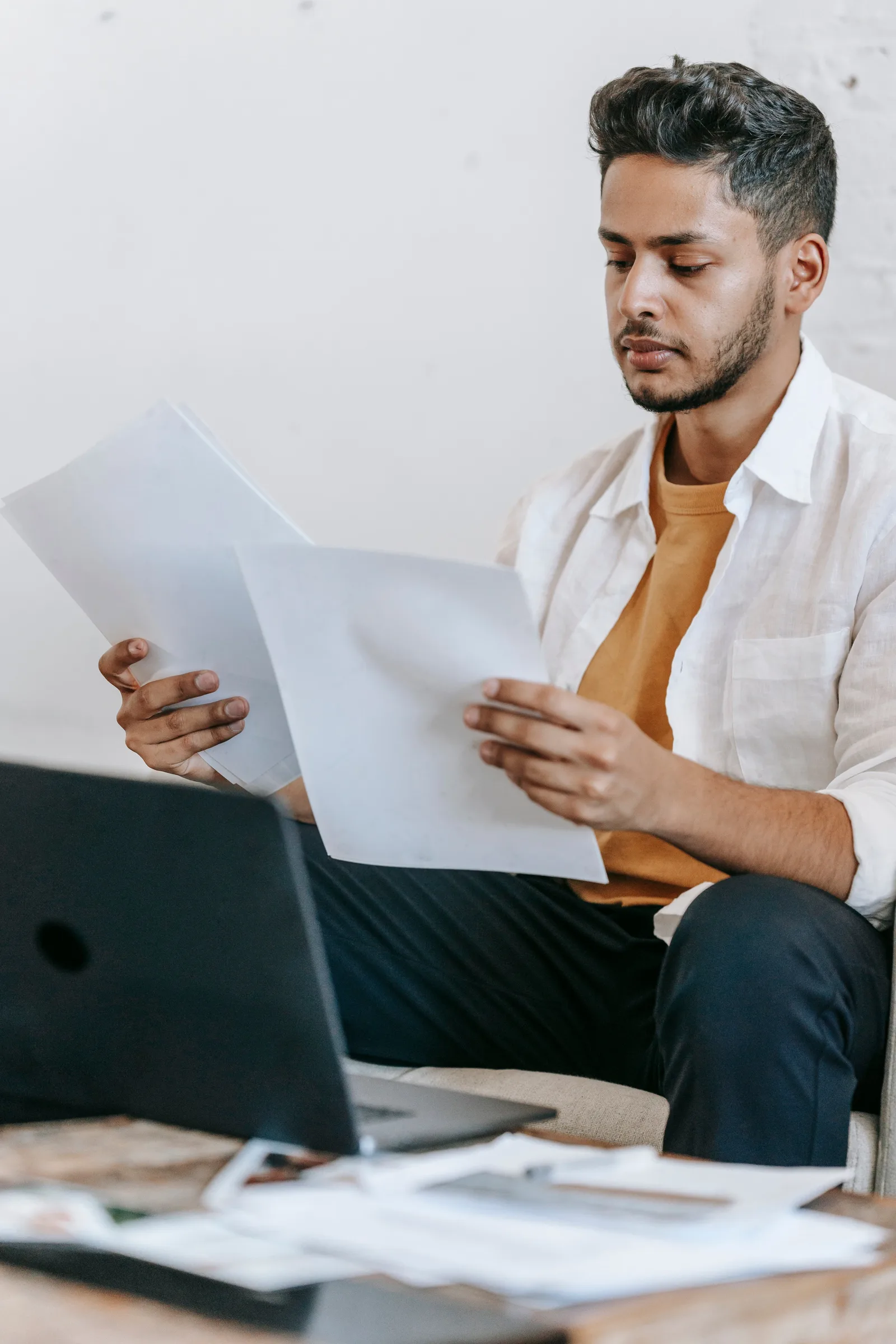 Serious young bearded Hispanic male journalist reviewing article on paper while working at table with laptop at workplace