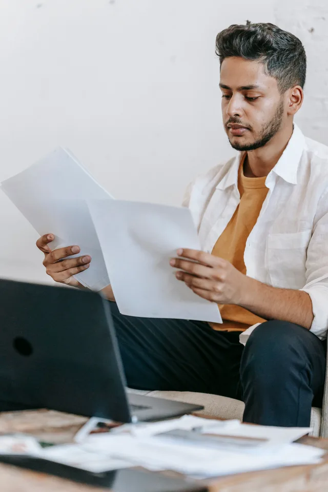 Serious young bearded Hispanic male journalist reviewing article on paper while working at table with laptop at workplace. Avviare un'impresa in Italia: adempimenti, costi e iscrizioni obbligatorie